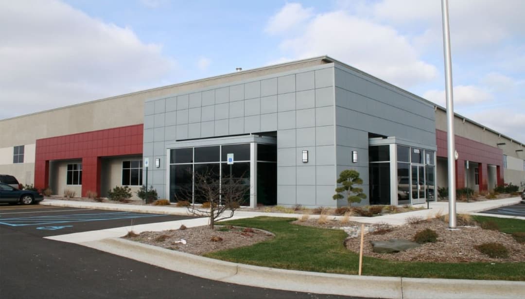 Single-story grey and red industrial and office building with a flagpole out front, under a partly cloudy sky