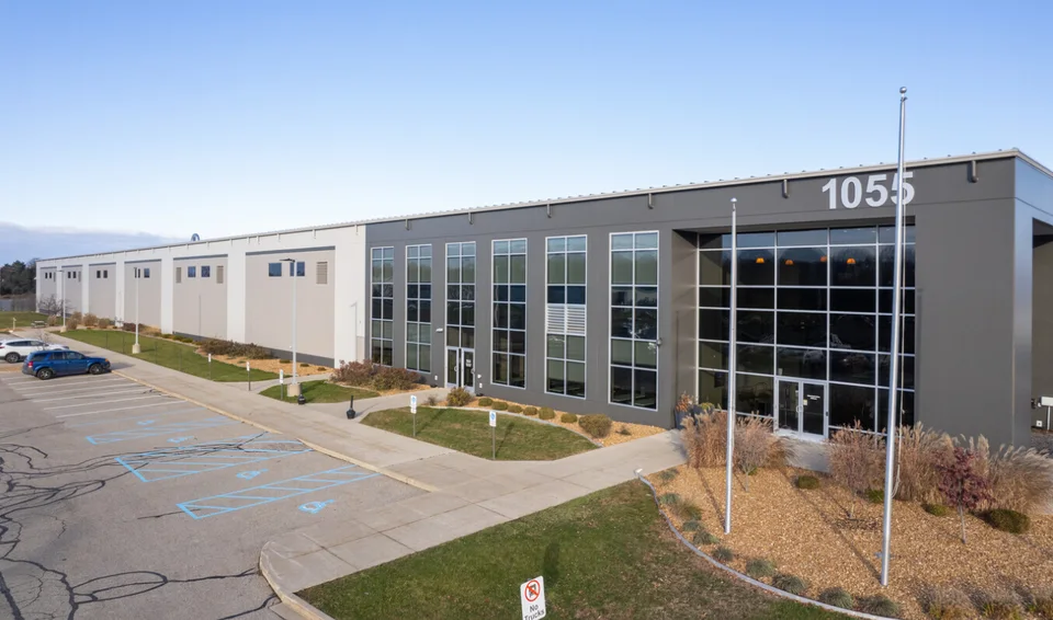 The two-story office entrance of a single-tenant industrial building, its gray metal facade carrying the numerals 1055 above a glazed curtain wall, with the long warehouse running off to the left and an empty parking lot in front.