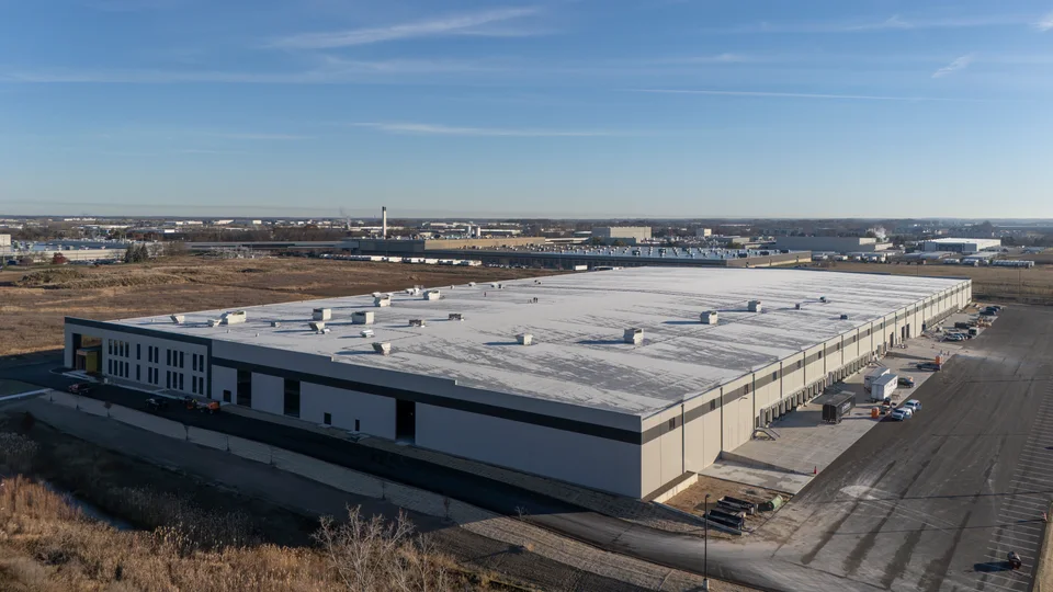 Aerial view of the 60th Street Warehouse, a large white distribution building with a flat roof, a row of dock doors and a paved truck court along one side, and other industrial buildings on the horizon.