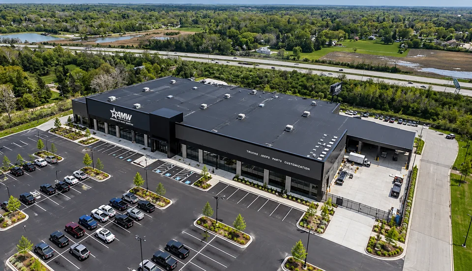 Aerial view of a long black-clad commercial building with AMW signage on the front elevation and lettering reading trucks, jeeps, parts, customization, a filled parking lot in front and a highway beyond.