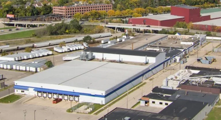 Aerial view of 320 Hall Street SW, a long single-story industrial building with a white roof, a blue band along its walls and a row of loading docks, with rail sidings, parked trailers and an elevated highway behind it.
