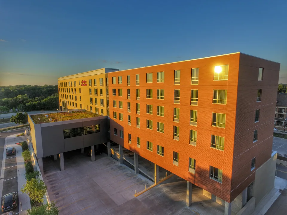 A six-story hotel at dusk, faced in orange brick and yellow panel, with a planted green roof over its lower wing and parking beneath the upper floors.